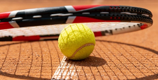 Une balle de tennis repose sur un court en terre battue orange, projetant une ombre, avec une raquette de tennis inclinée au-dessus. La scène dégage une impression d'action et de sport