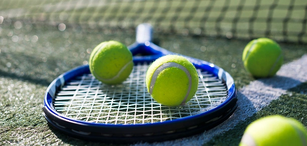 Une raquette de tennis bleue repose sur un court vert, entourée de plusieurs balles de tennis jaune vif. Un filet est visible dans l'arrière-plan flou